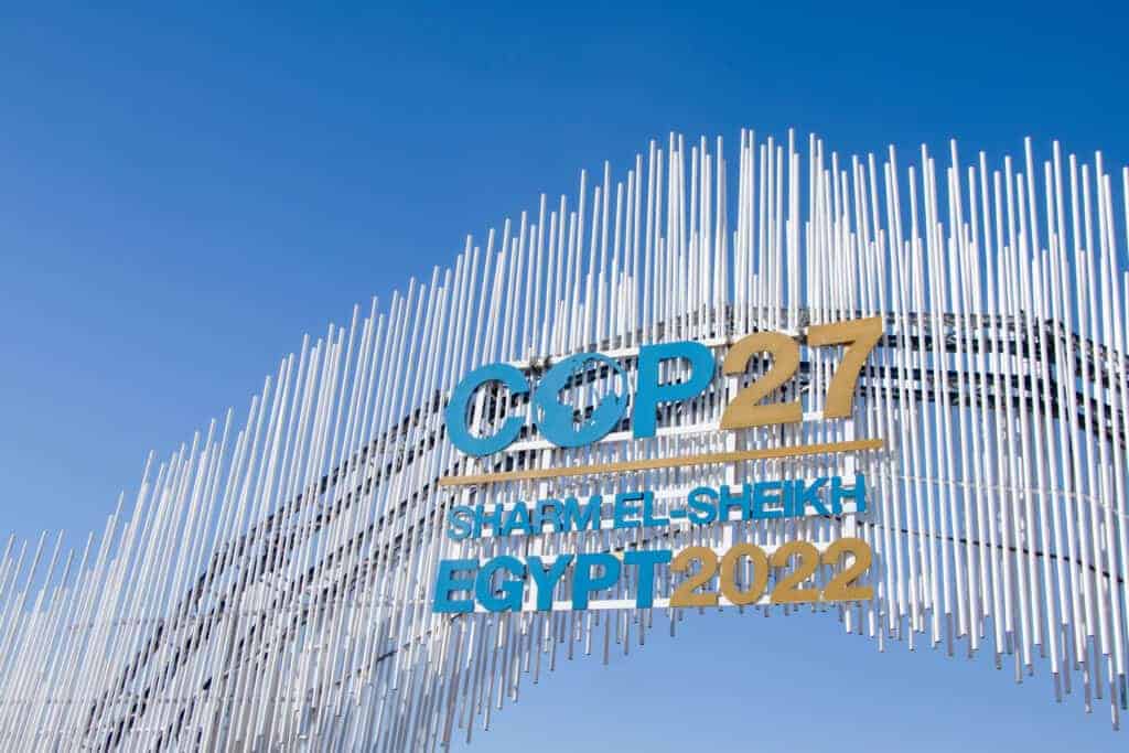 A large outdoor sign for COP27 Sharm El-Sheikh Egypt 2022, featuring blue and gold text on a structure of white vertical rods against a blue sky.