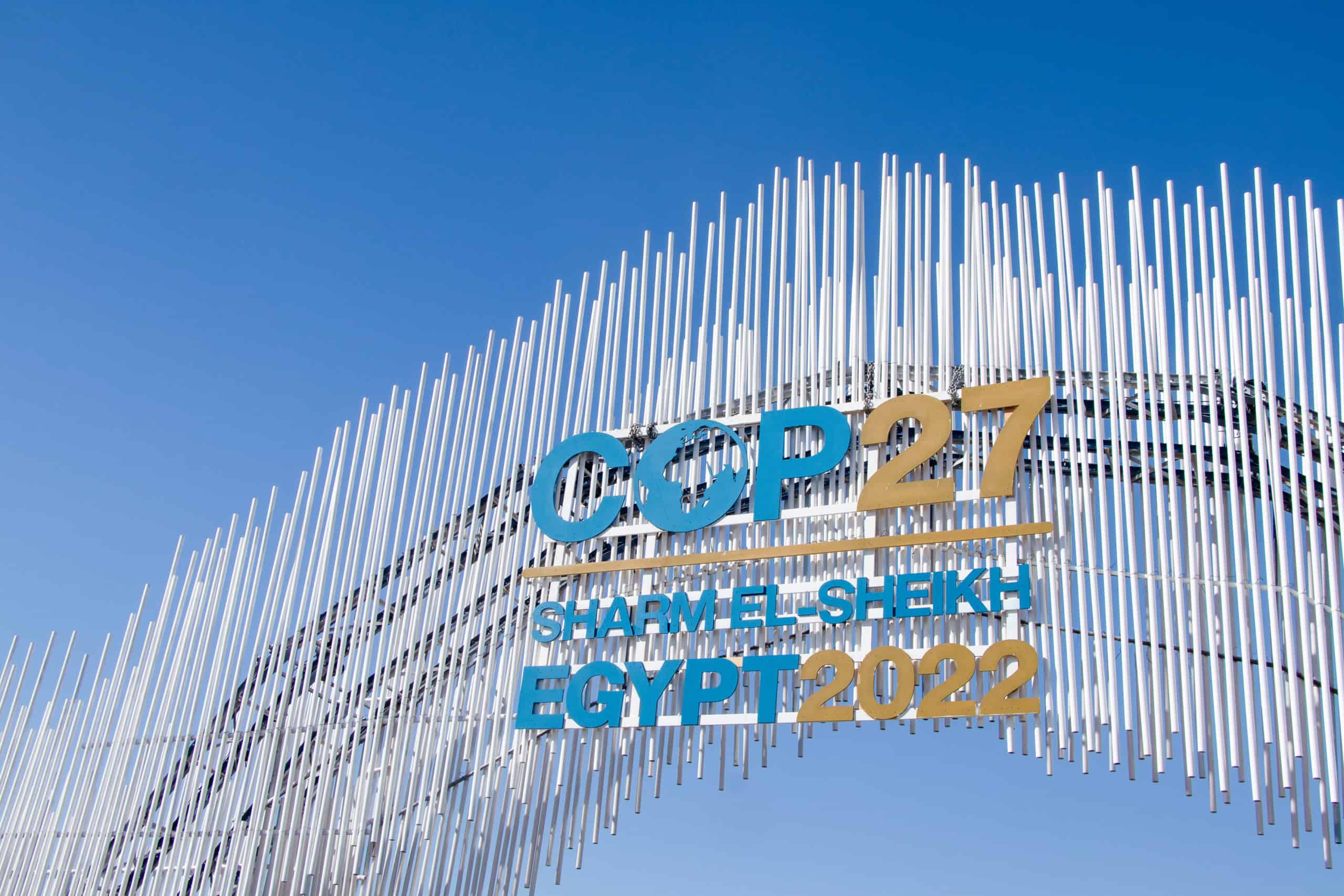 A large outdoor sign for COP27 Sharm El-Sheikh Egypt 2022, featuring blue and gold text on a structure of white vertical rods against a blue sky.