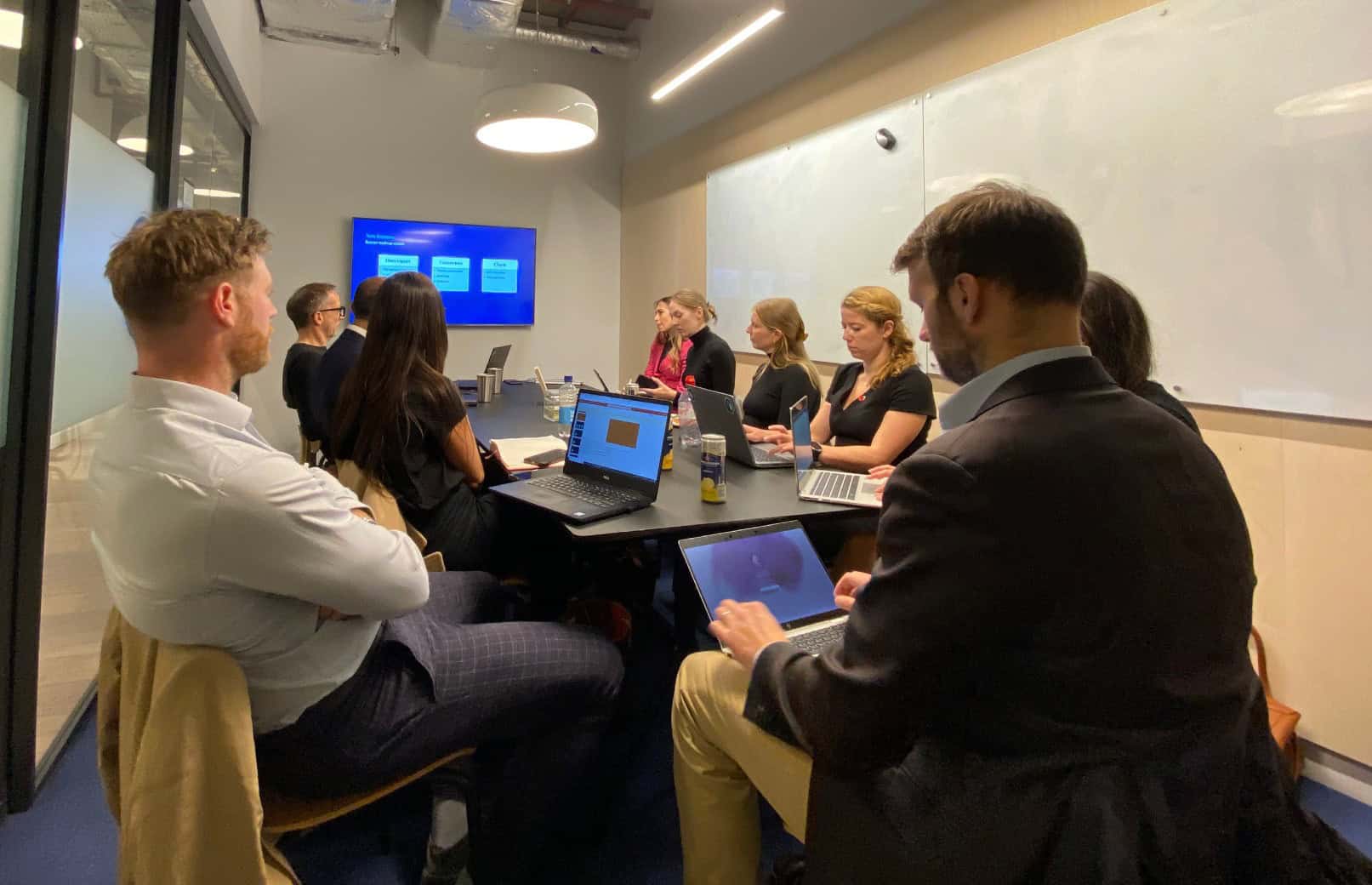 A group of people seated around a table in a meeting room, looking at a presentation on a screen and working on laptops.