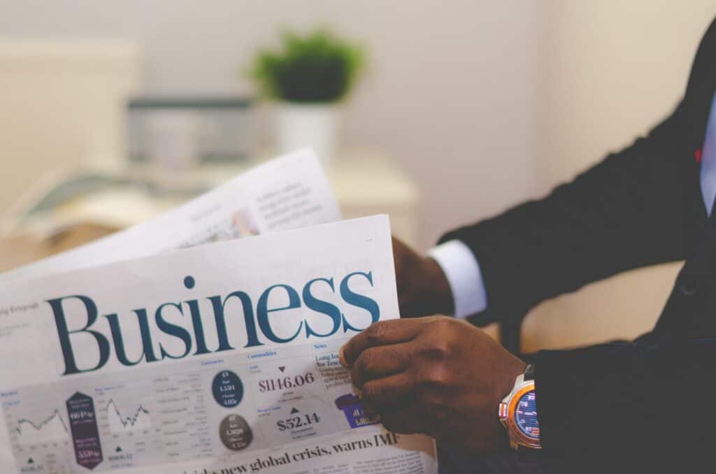 A person in a suit holds a newspaper open to the 'Business' section, with financial figures and headlines visible.