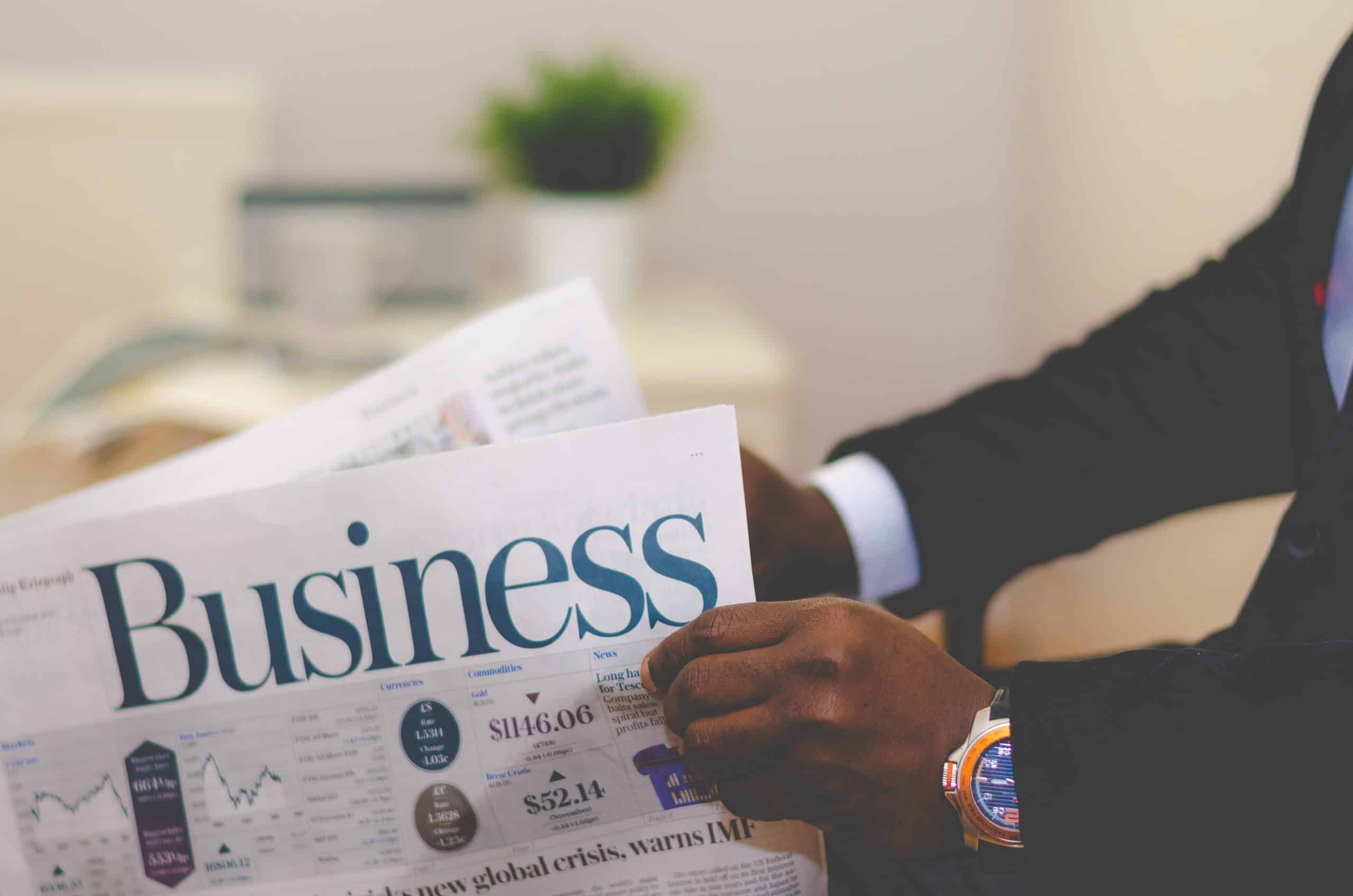 A person in a suit holds a newspaper open to the 'Business' section, with financial figures and headlines visible.
