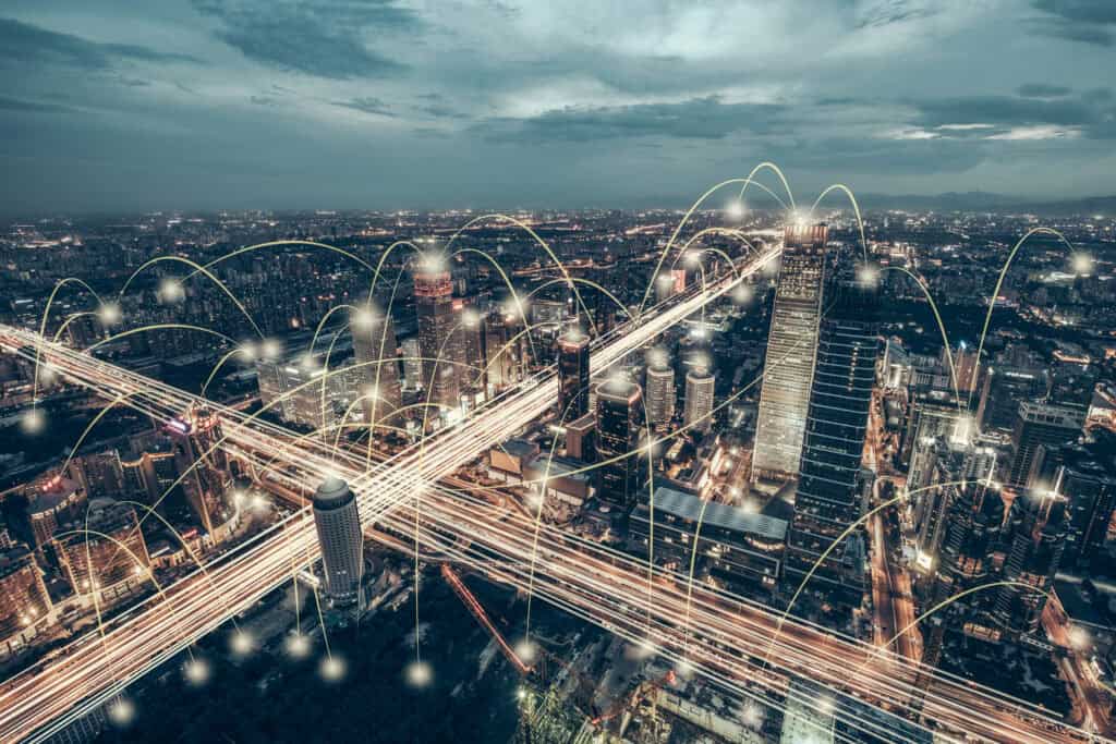 Aerial view of a city at night with glowing light trails on roads and arcing lines connecting buildings, symbolizing a network.