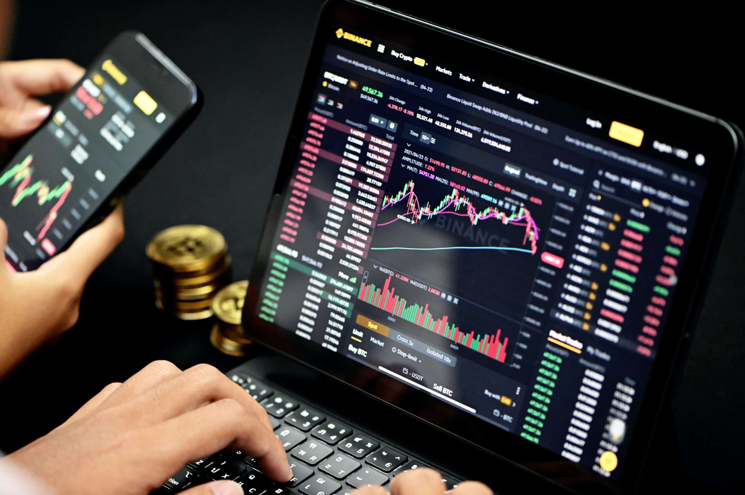 A person's hands using a laptop and smartphone to monitor cryptocurrency trading charts on Binance, with gold-colored coins in the background.