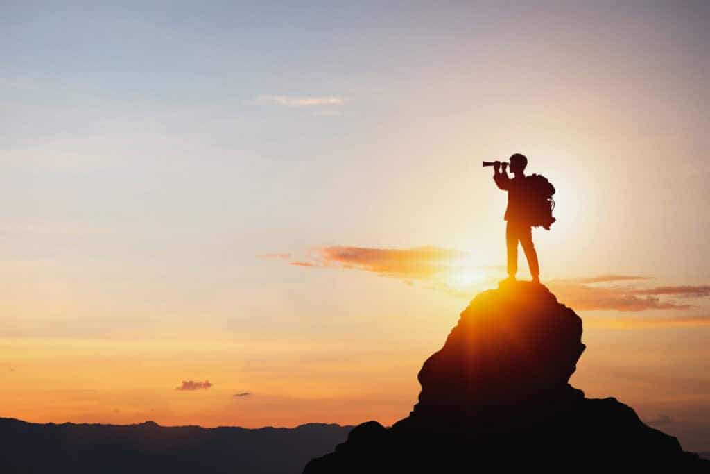 Silhouette of a person with a backpack standing on a mountain peak, looking through a monocular at a bright sunrise or sunset.