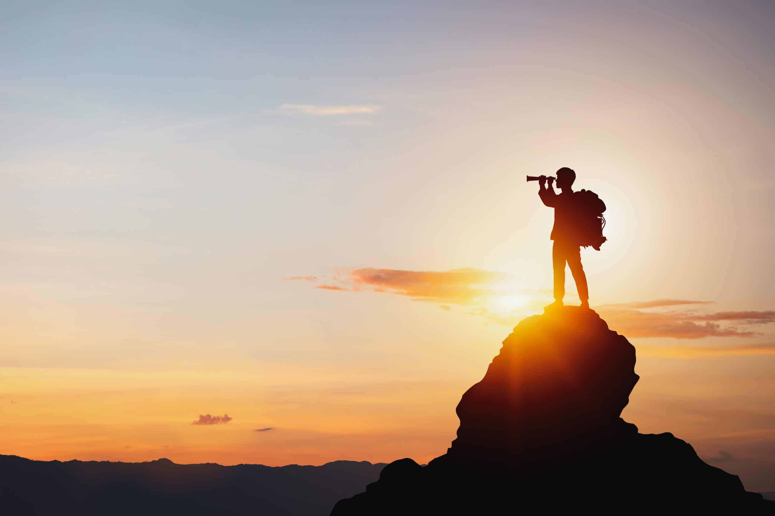 Silhouette of a person with a backpack standing on a mountain peak, looking through a monocular at a bright sunrise or sunset.