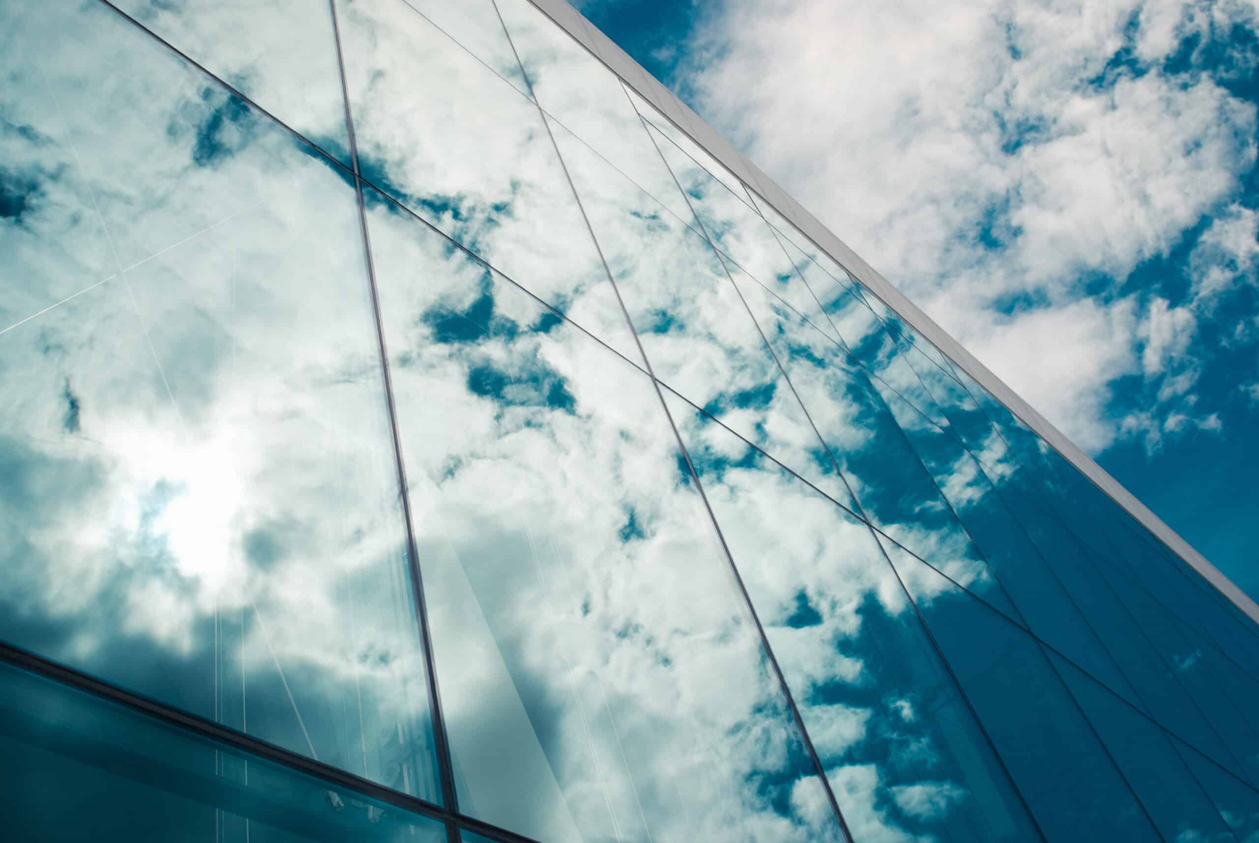 Close-up of a modern glass building facade reflecting a bright, cloudy blue sky with a sun glare.