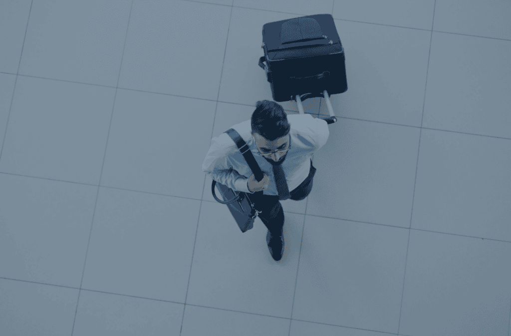 Top-down view of a man in a light blue shirt and dark tie, pulling a black suitcase on a tiled floor.