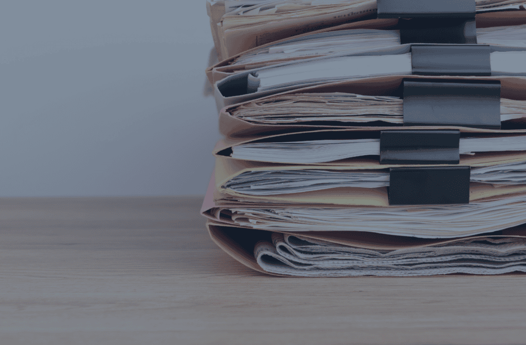 A tall stack of files and documents in various colored folders, secured with black binder clips, resting on a wooden surface.