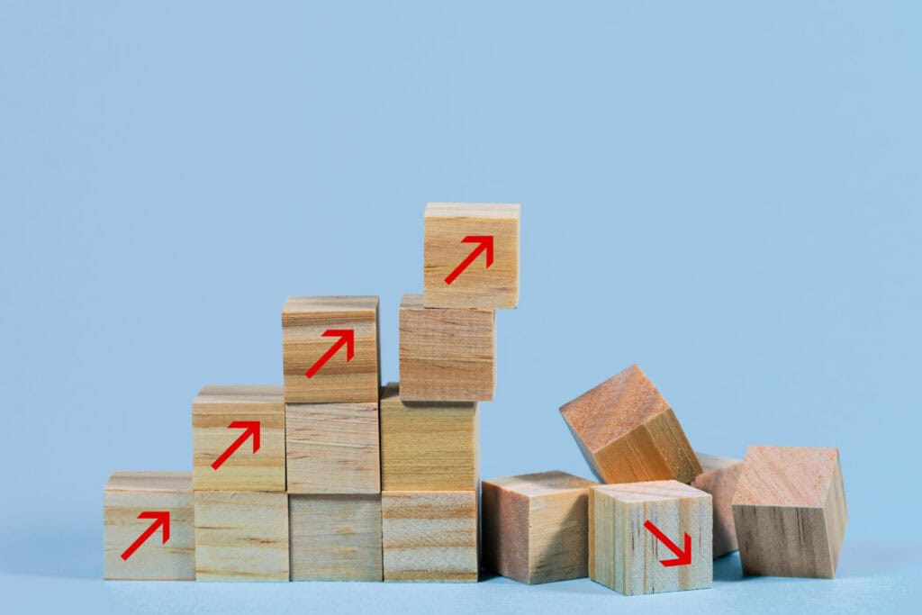 A staircase of wooden blocks with red upward arrows on the left, contrasting with scattered blocks on the right, one with a red downward arrow, all on a light blue background.