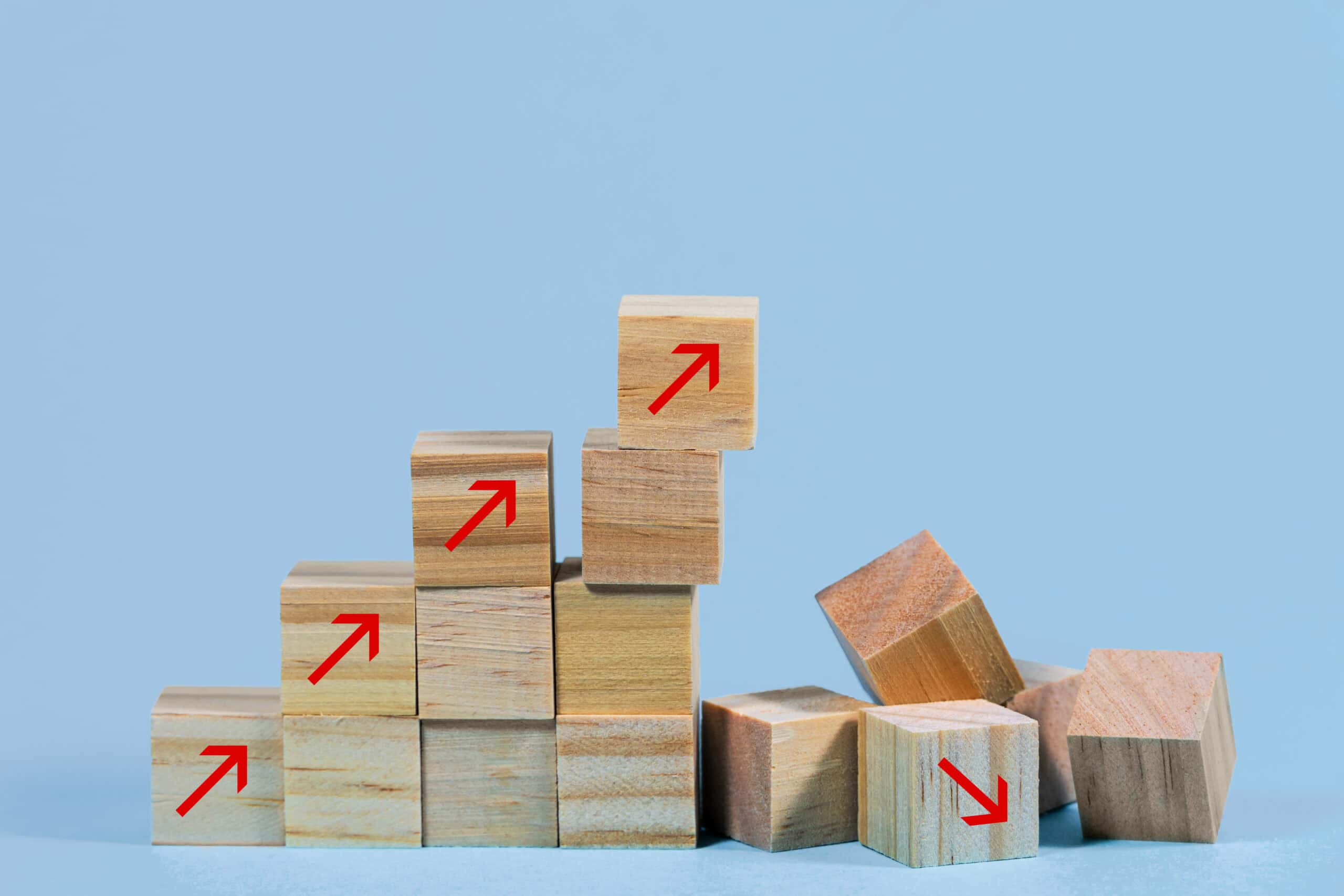 A staircase of wooden blocks with red upward arrows on the left, contrasting with scattered blocks on the right, one with a red downward arrow, all on a light blue background.