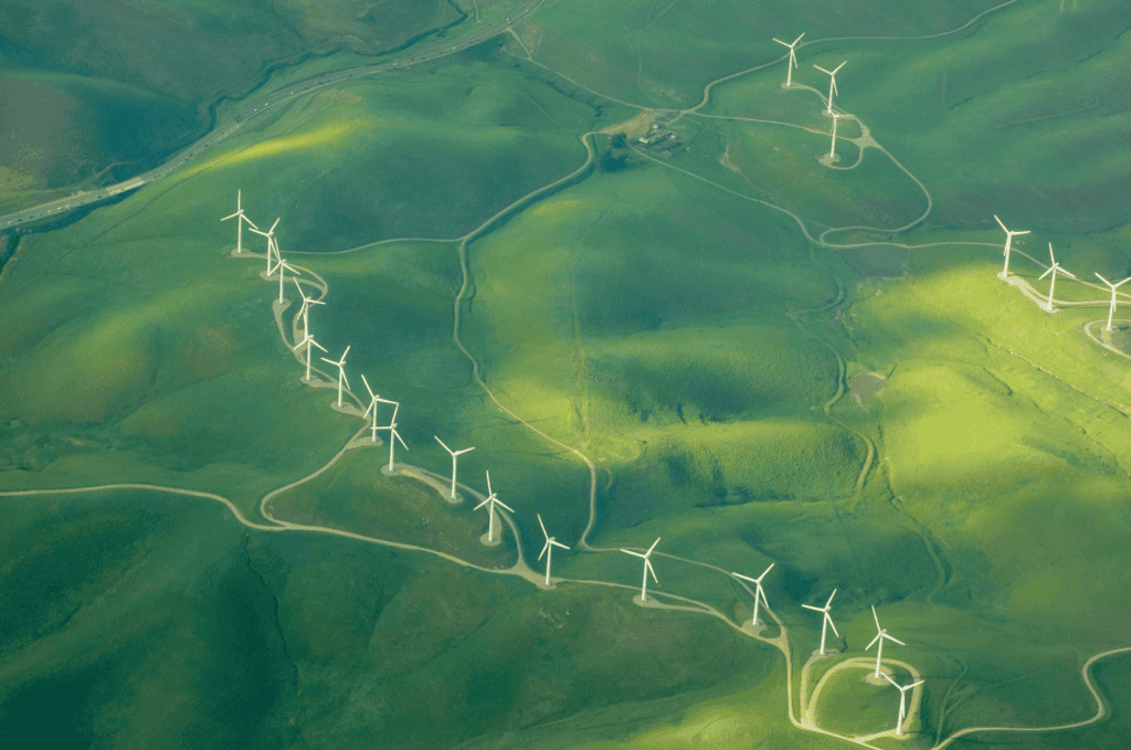 Aerial view of a wind farm with numerous white wind turbines spread across rolling green hills, connected by dirt roads. A highway is visible in the distance.