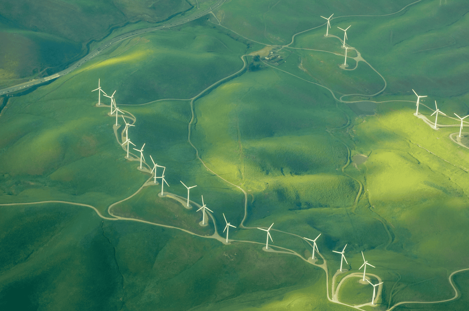 Aerial view of a wind farm with numerous white wind turbines spread across rolling green hills, connected by dirt roads. A highway is visible in the distance.