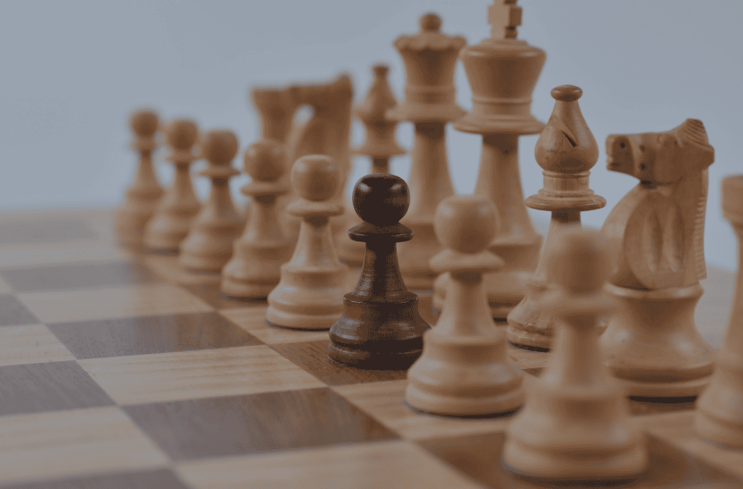 A dark brown chess pawn stands on a chessboard, surrounded by many light-colored chess pieces.