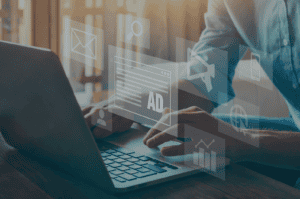 Close-up of hands typing on a laptop keyboard, with various translucent digital marketing icons overlaid, including email, search, display ads, mobile ads, and analytics.