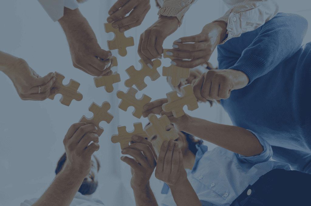 Multiple hands hold wooden puzzle pieces, seen from a low angle, symbolizing teamwork and collaboration.