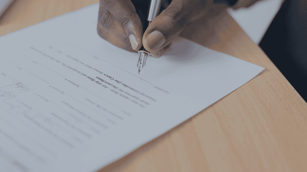 A dark-skinned hand holds a silver fountain pen, signing a white document on a wooden desk.