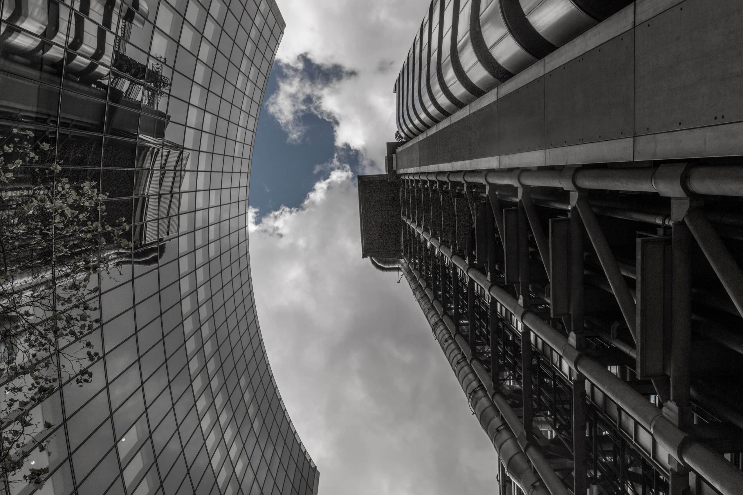 Low-angle shot of two tall buildings against a cloudy sky with a patch of blue. One building is curved glass, reflecting trees; the other is industrial with exposed pipes.