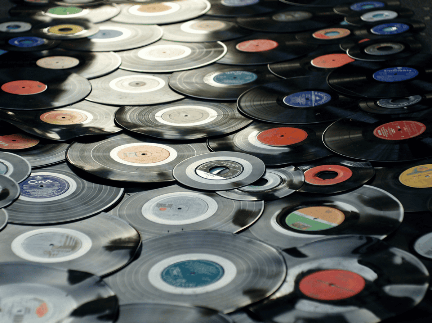 A close-up, overhead view of numerous black vinyl records and 45s scattered and overlapping on a dark surface. The records have colorful labels in various hues.