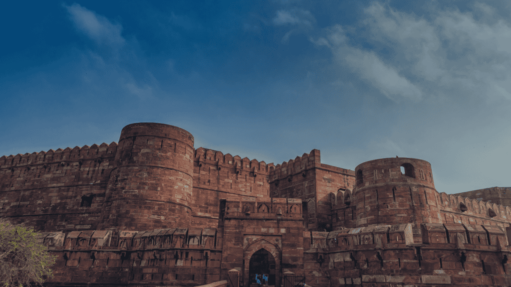 A large, red sandstone fort with high walls, circular bastions, and an arched entrance under a blue sky with white clouds.