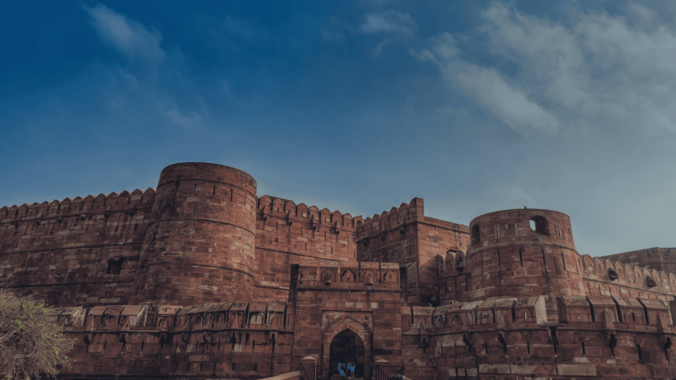 A large, red sandstone fort with high walls, circular bastions, and an arched entrance under a blue sky with white clouds.
