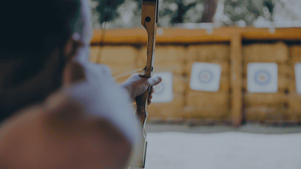 A person seen from behind, holding a wooden bow and aiming at blurred archery targets in the background.