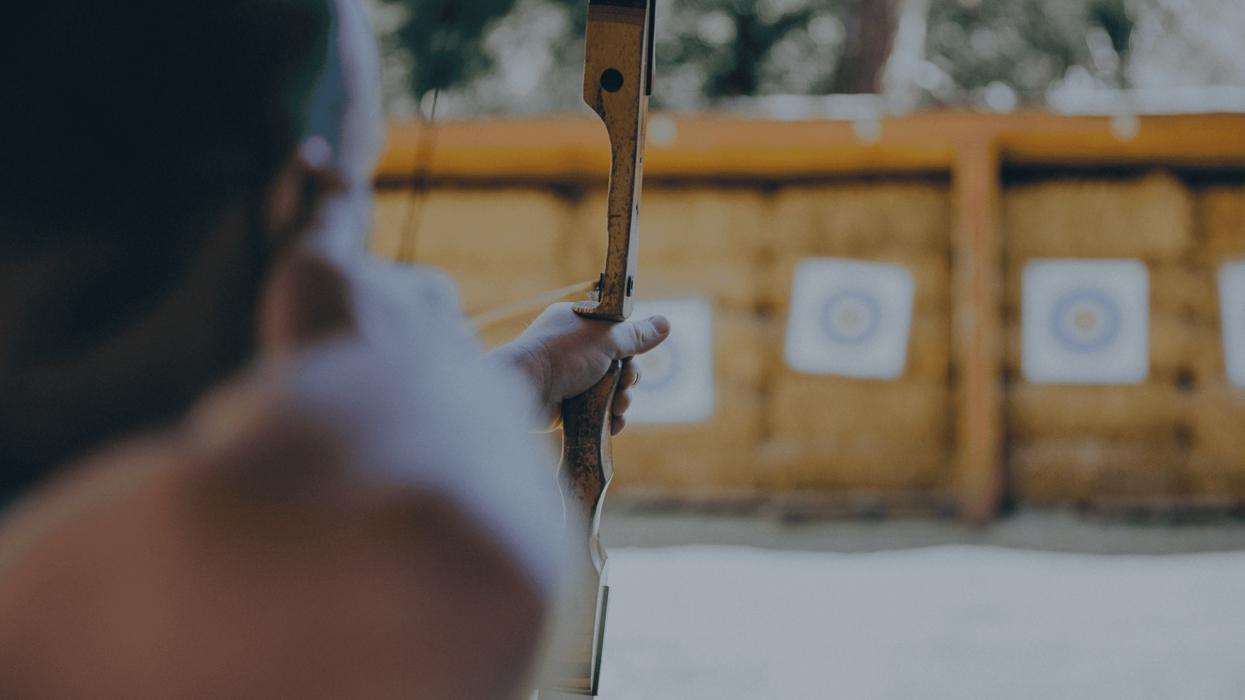 A person seen from behind, holding a wooden bow and aiming at blurred archery targets in the background.