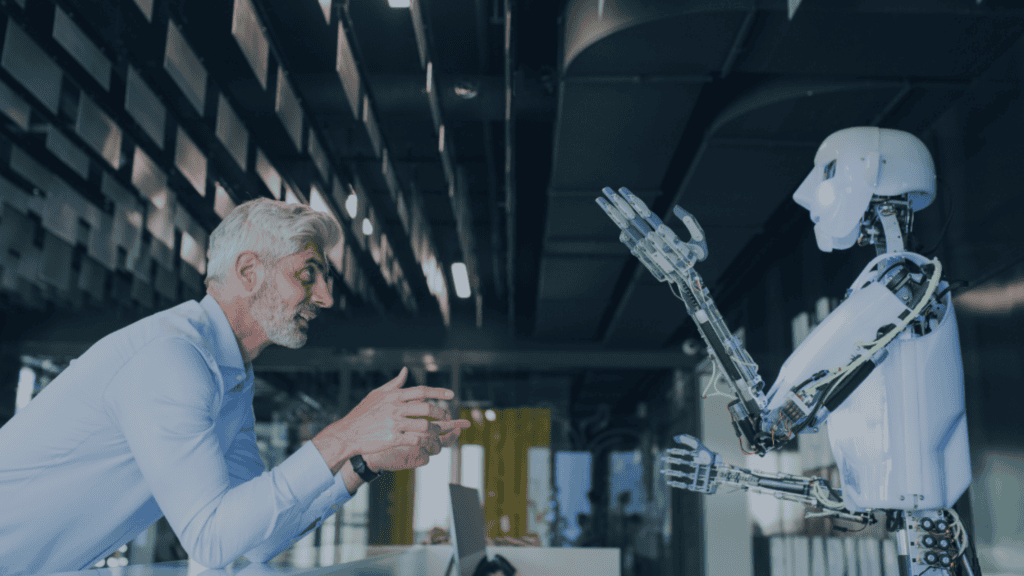 A man with gray hair and beard in a blue shirt talks to a white humanoid robot with exposed wiring, both gesturing, in a modern office setting.