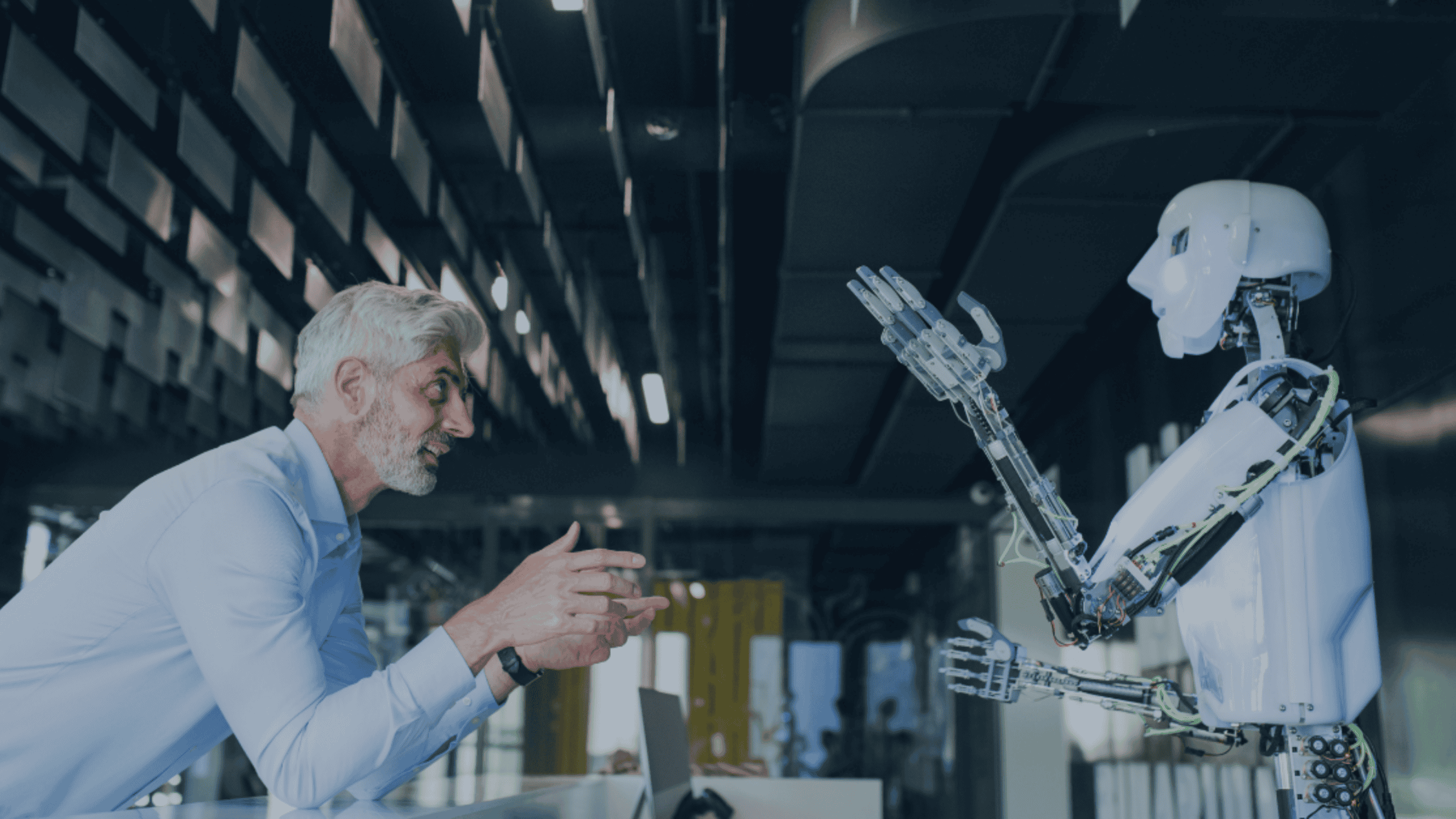 A man with gray hair and beard in a blue shirt talks to a white humanoid robot with exposed wiring, both gesturing, in a modern office setting.
