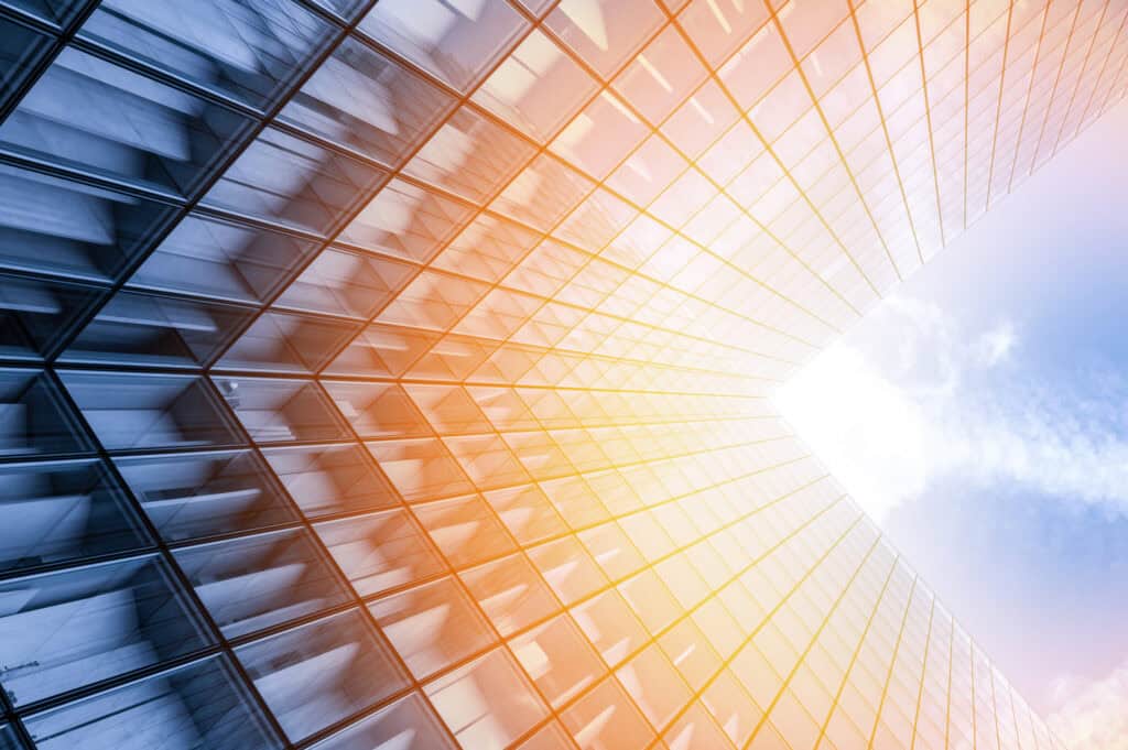 Low-angle view of a modern glass skyscraper reflecting a blue sky and bright orange-yellow sunlight, creating a vibrant gradient.