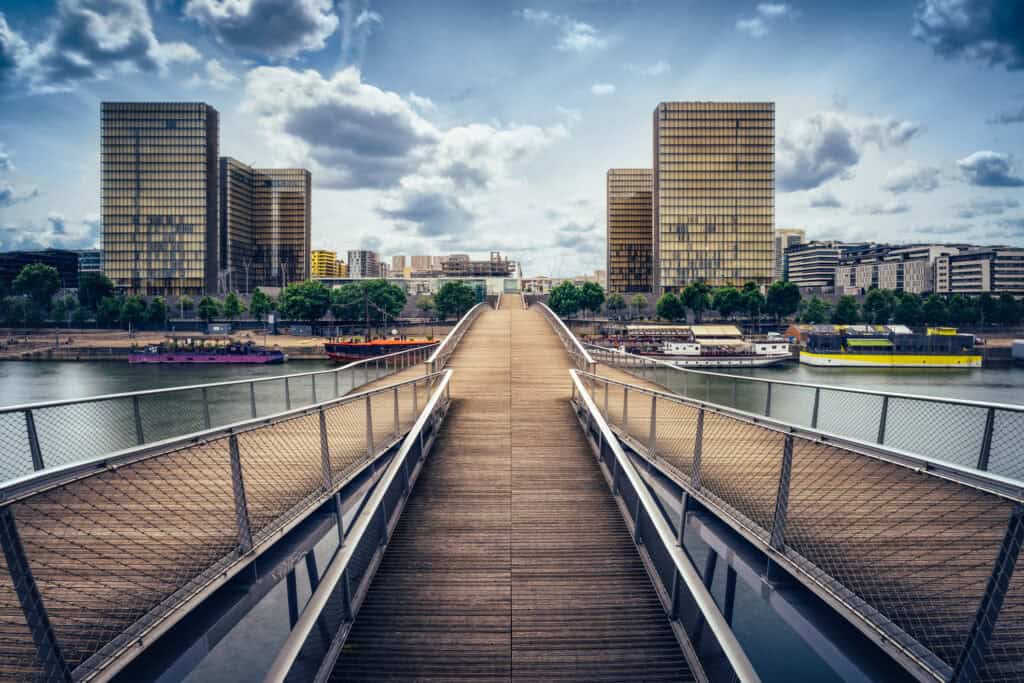 A pedestrian bridge with wooden planks and metal railings extends over a river towards modern, tall buildings under a cloudy sky.