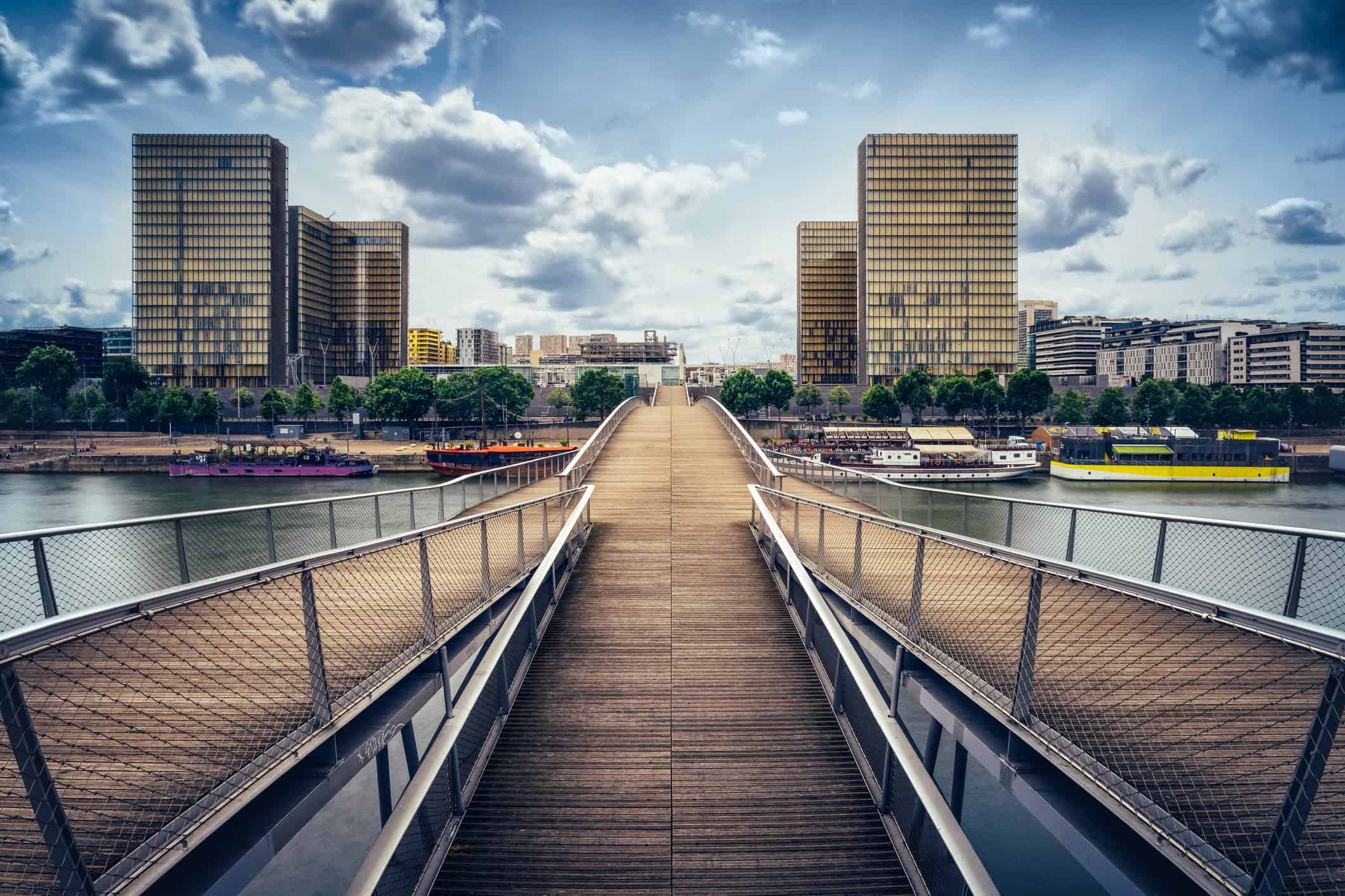 A pedestrian bridge with wooden planks and metal railings extends over a river towards modern, tall buildings under a cloudy sky.