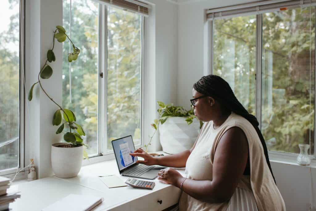 A Black woman with braided hair and glasses works on a laptop at a white desk by a large window, surrounded by potted plants.