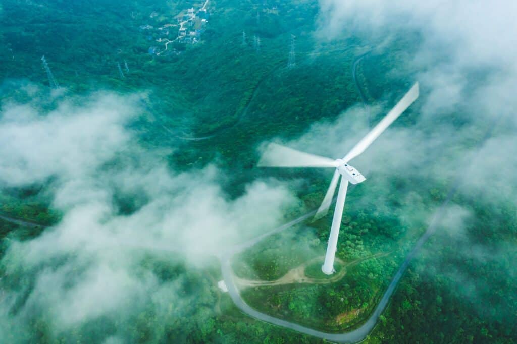 Aerial view of a white wind turbine with blurred blades, standing on a green, forested mountain. White clouds or fog partially cover the landscape, with roads, buildings, and power lines visible in the distance.