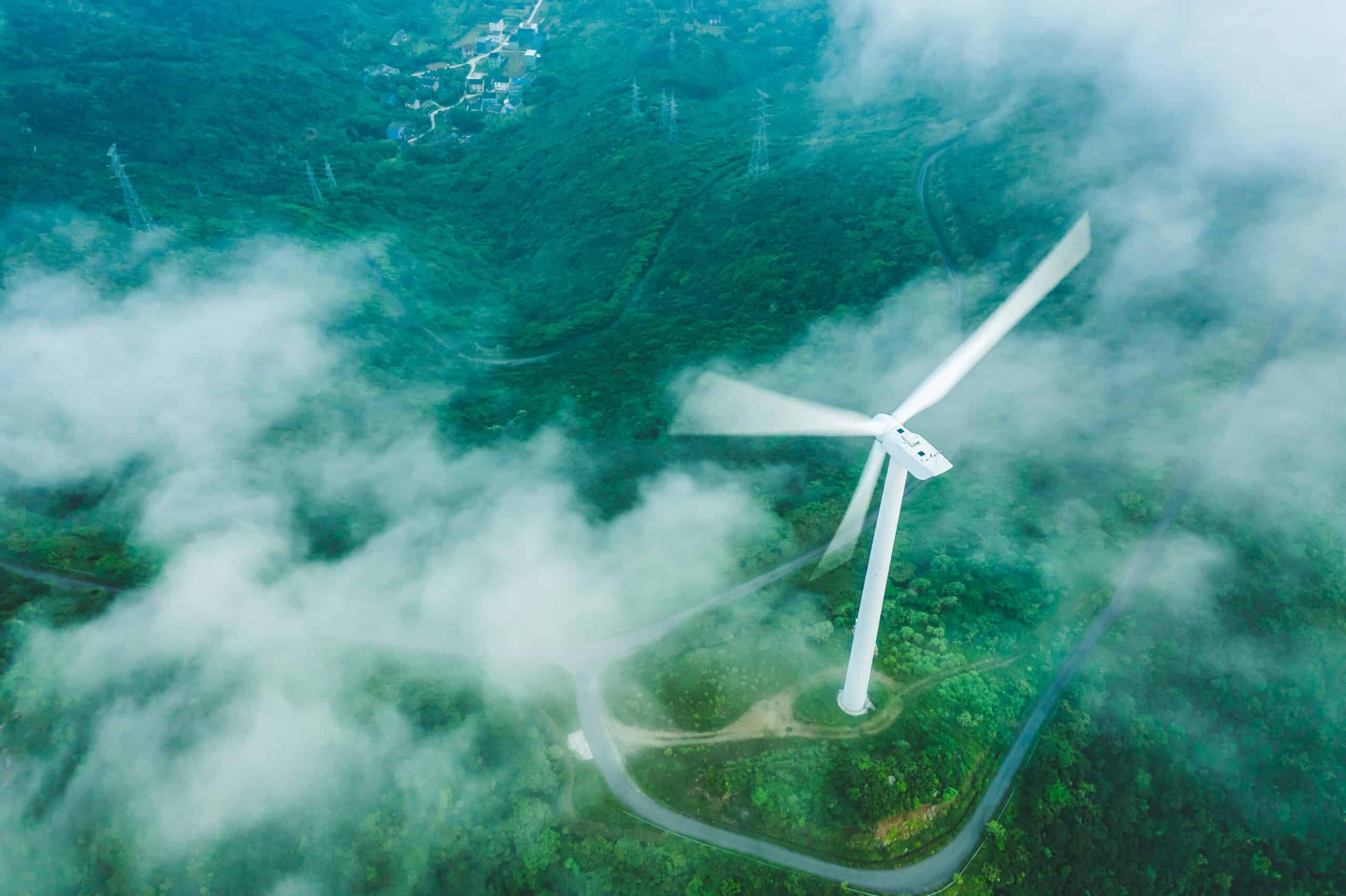 Aerial view of a white wind turbine with blurred blades, standing on a green, forested mountain. White clouds or fog partially cover the landscape, with roads, buildings, and power lines visible in the distance.
