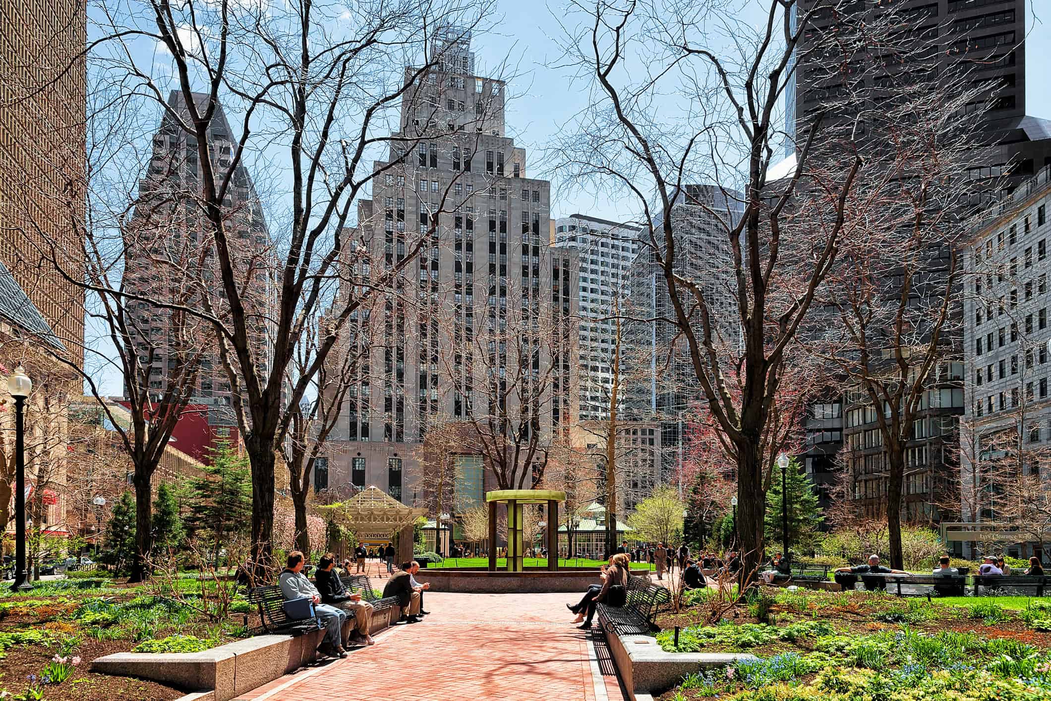 An urban park with bare trees, green landscaping, and people on benches, surrounded by tall city buildings under a blue sky.