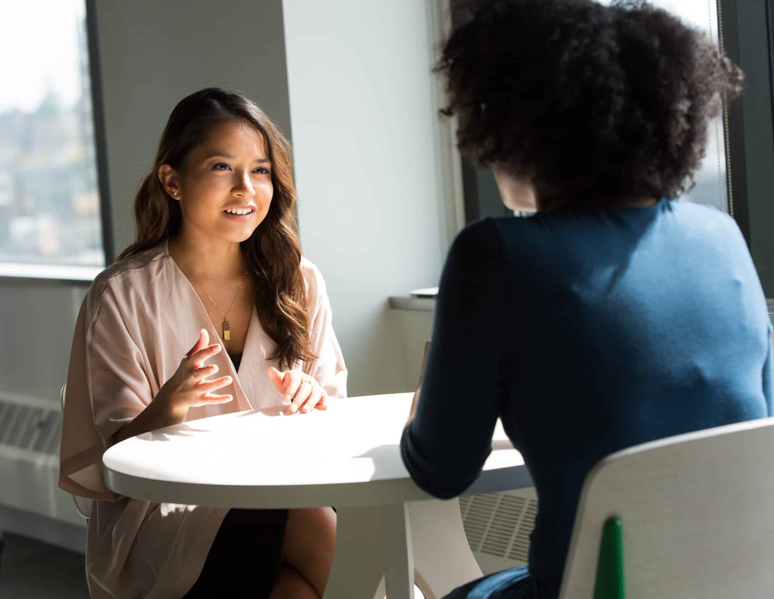 Two women sitting at a white table, one facing the camera and gesturing while talking, the other seen from behind, both appearing to be in conversation.