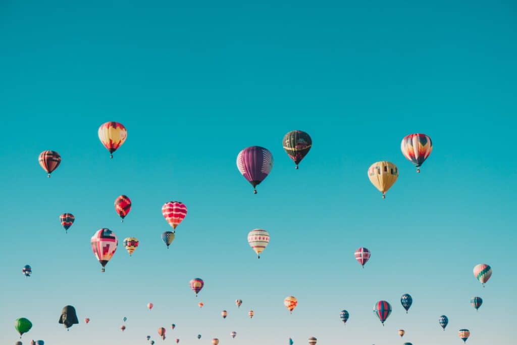 Many colorful hot air balloons, including one shaped like Darth Vader's helmet, float across a clear blue sky.