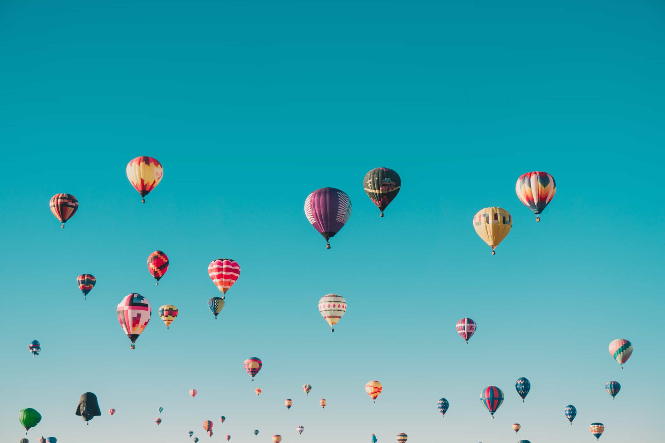 Many colorful hot air balloons, including one shaped like Darth Vader's helmet, float across a clear blue sky.