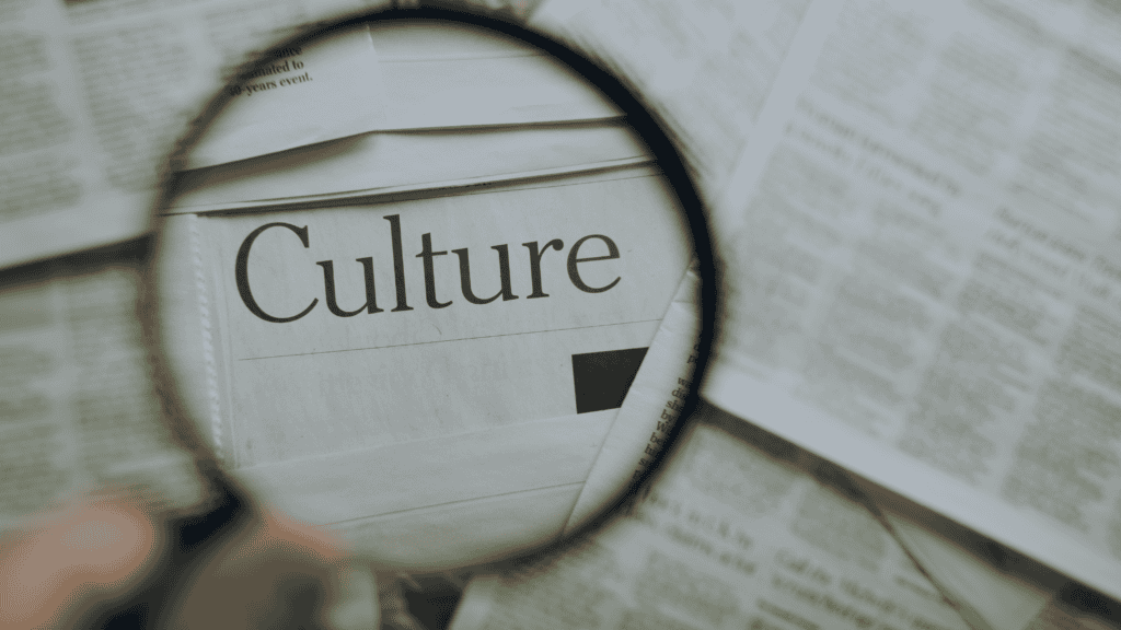 A magnifying glass held by a hand over a newspaper, clearly showing the word 'Culture' magnified within the lens. Surrounding text is blurred.