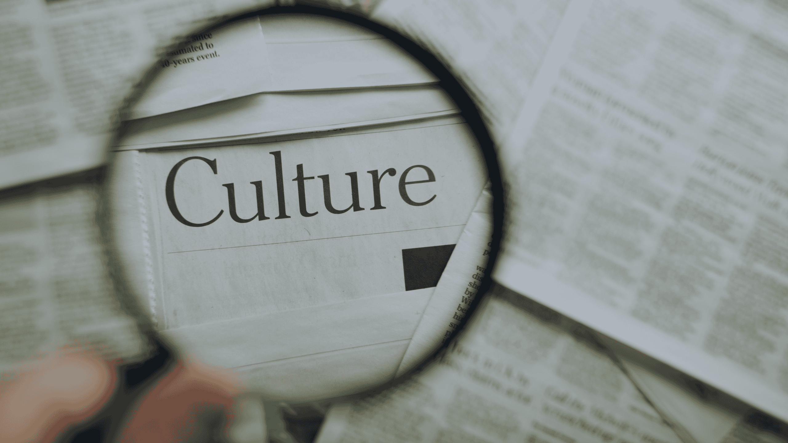 A magnifying glass held by a hand over a newspaper, clearly showing the word 'Culture' magnified within the lens. Surrounding text is blurred.