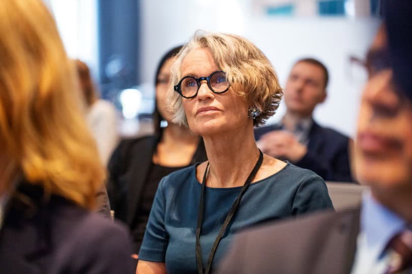 A woman with short gray hair and black-framed glasses looks up with a focused expression, wearing a teal dress and a lanyard, in a blurred conference setting.