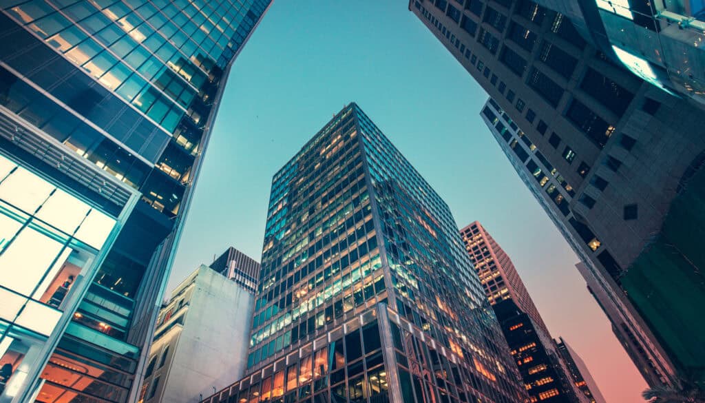 A low-angle view of multiple glass skyscrapers with illuminated windows against a twilight sky.