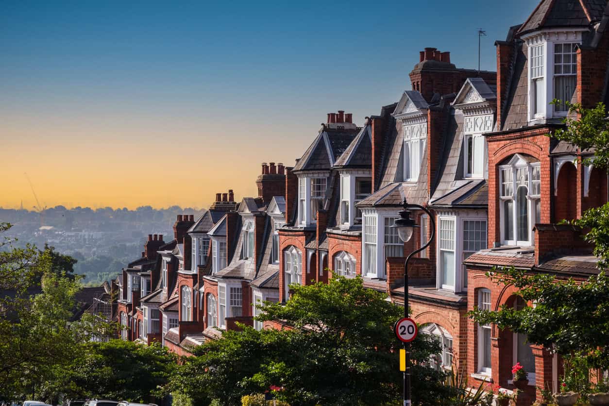 A row of red brick terraced houses with bay windows and slate roofs, overlooking a distant hazy city skyline under a gradient orange and blue sky.