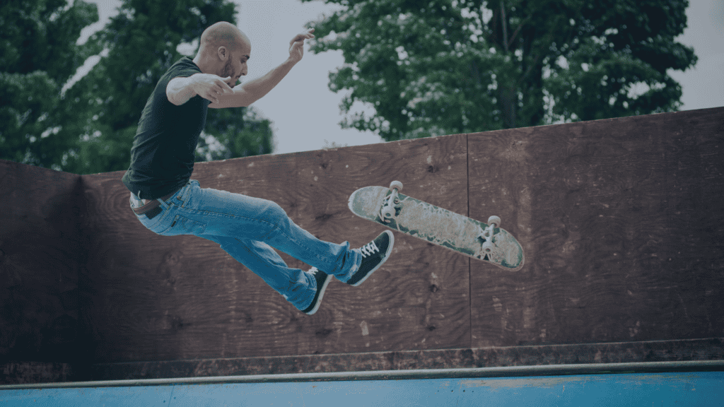 A bald man in a black t-shirt and blue jeans is mid-air, performing a skateboarding trick over a wooden ramp, with his skateboard floating nearby.