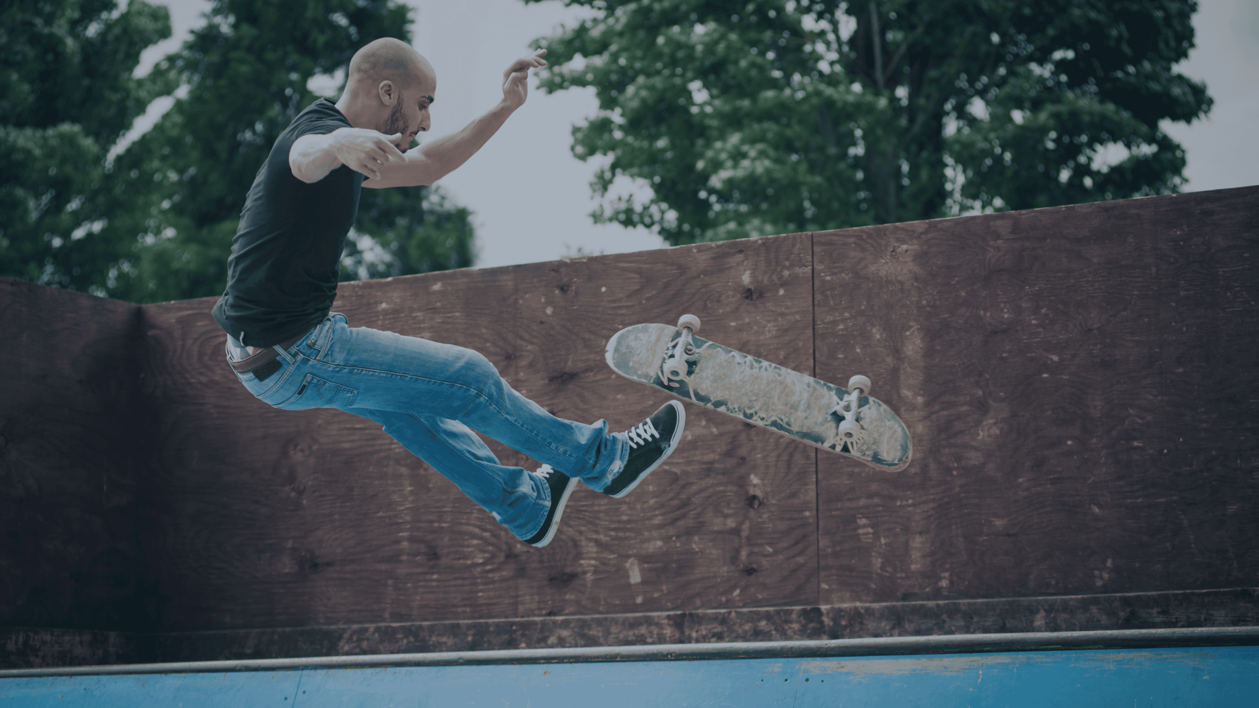 A bald man in a black t-shirt and blue jeans is mid-air, performing a skateboarding trick over a wooden ramp, with his skateboard floating nearby.