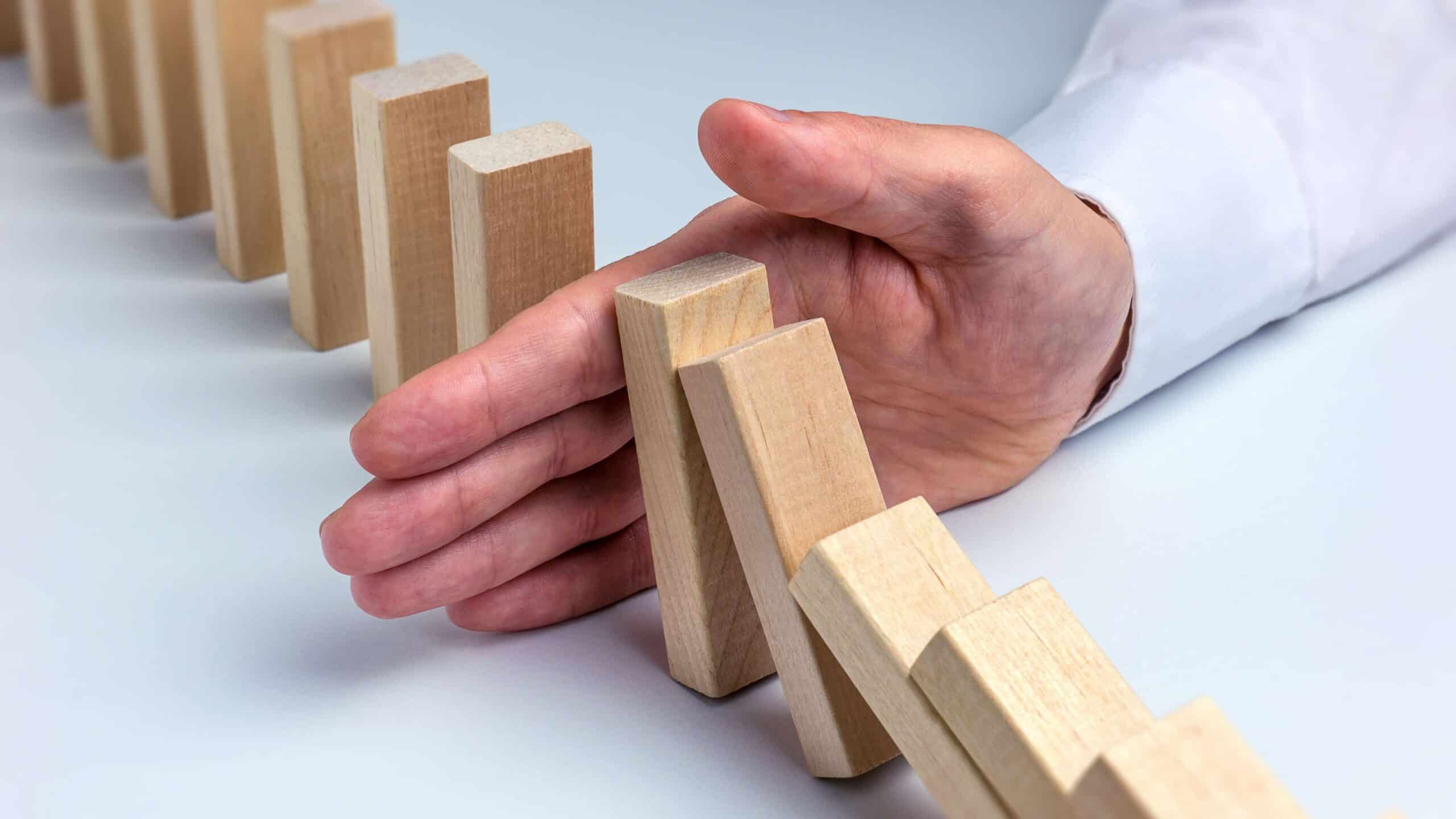 A person's hand in a white shirt cuff stops a line of falling wooden domino blocks.