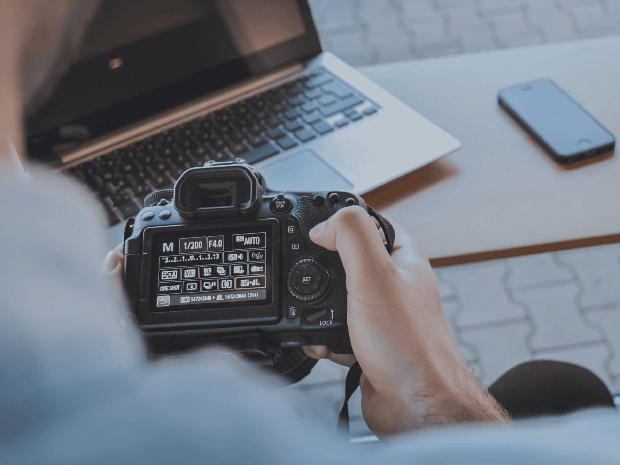 A person holds a black DSLR camera, its screen displaying various settings, next to an open laptop and a smartphone on a table.