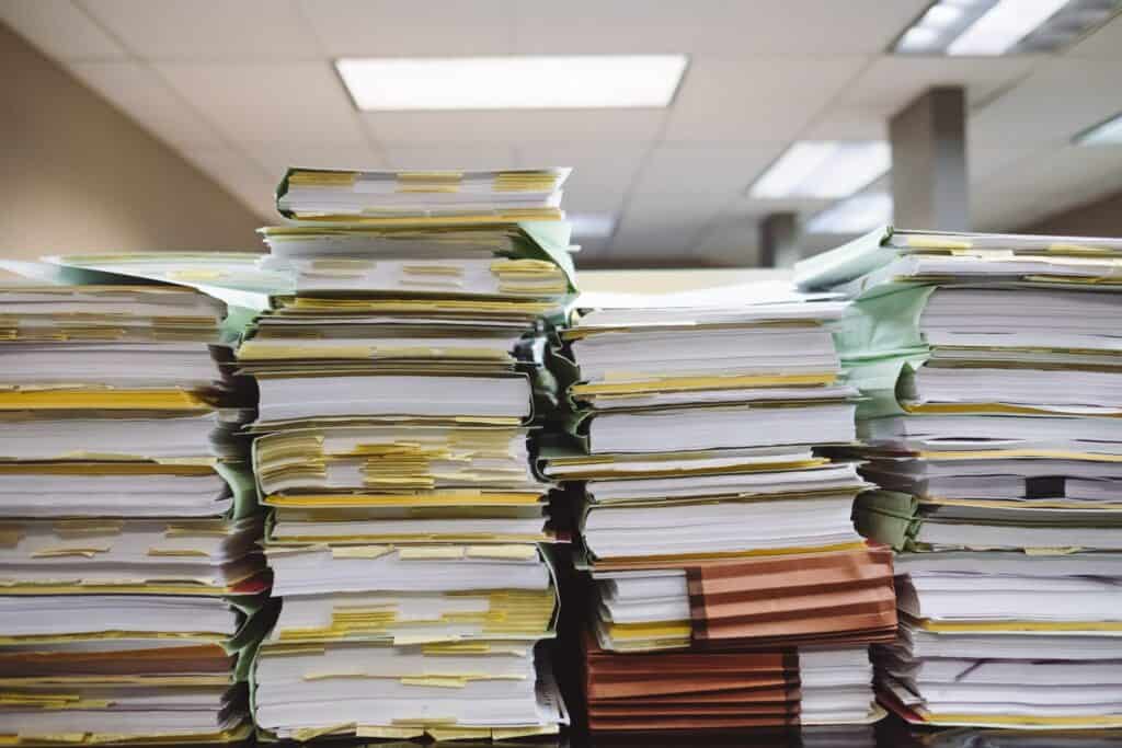 Close-up of numerous tall stacks of paper documents and files in green, yellow, and brown folders, with sticky notes, in an office setting.