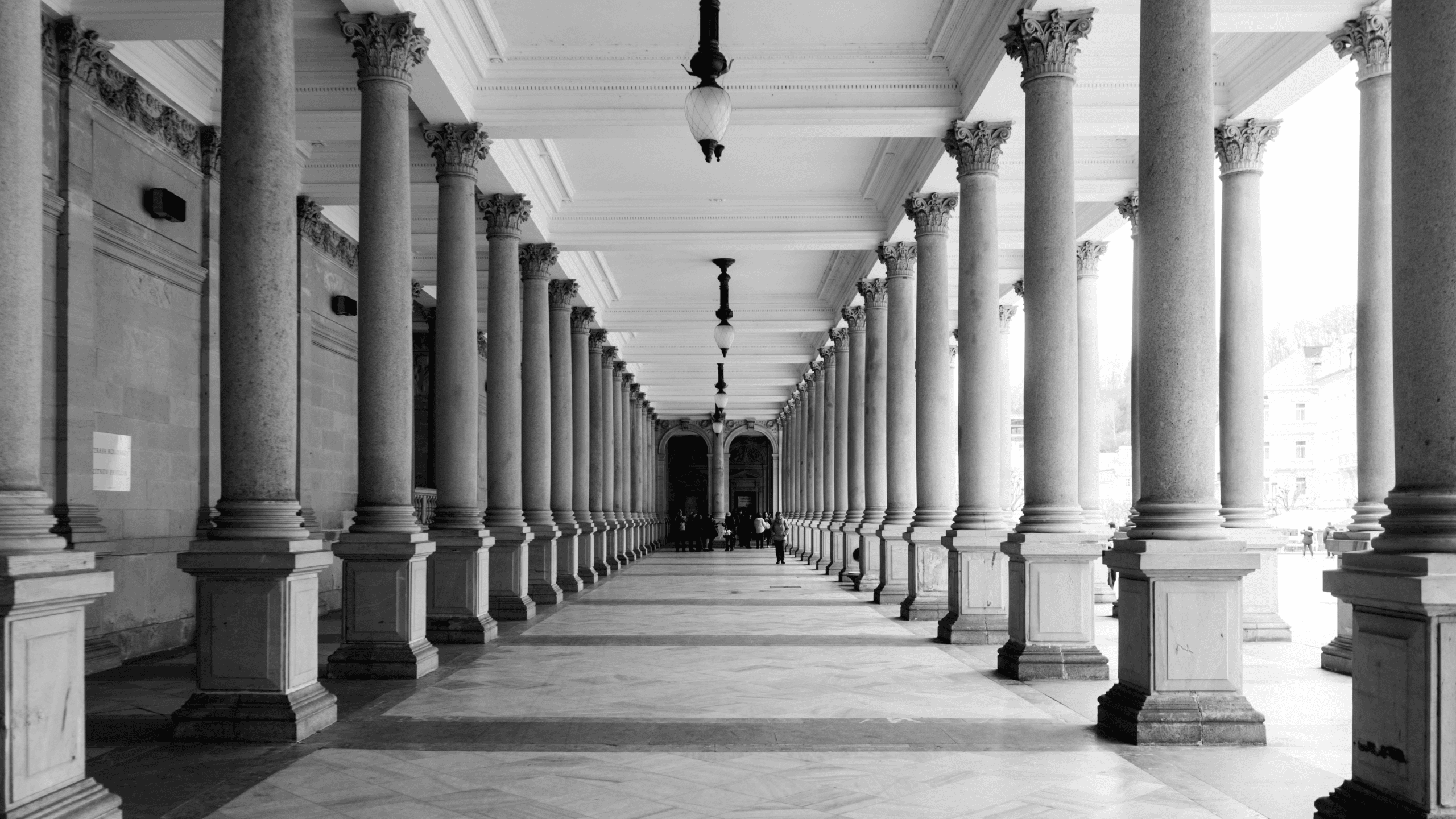 Black and white photo of a long, symmetrical colonnade with numerous fluted columns and ornate capitals, leading to a vanishing point with people in the distance.