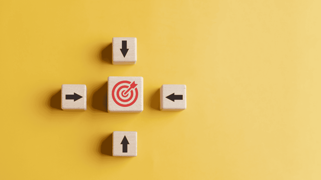 Five wooden blocks on a yellow background. A central block shows a red target with an arrow, surrounded by four blocks with black arrows pointing towards the center.